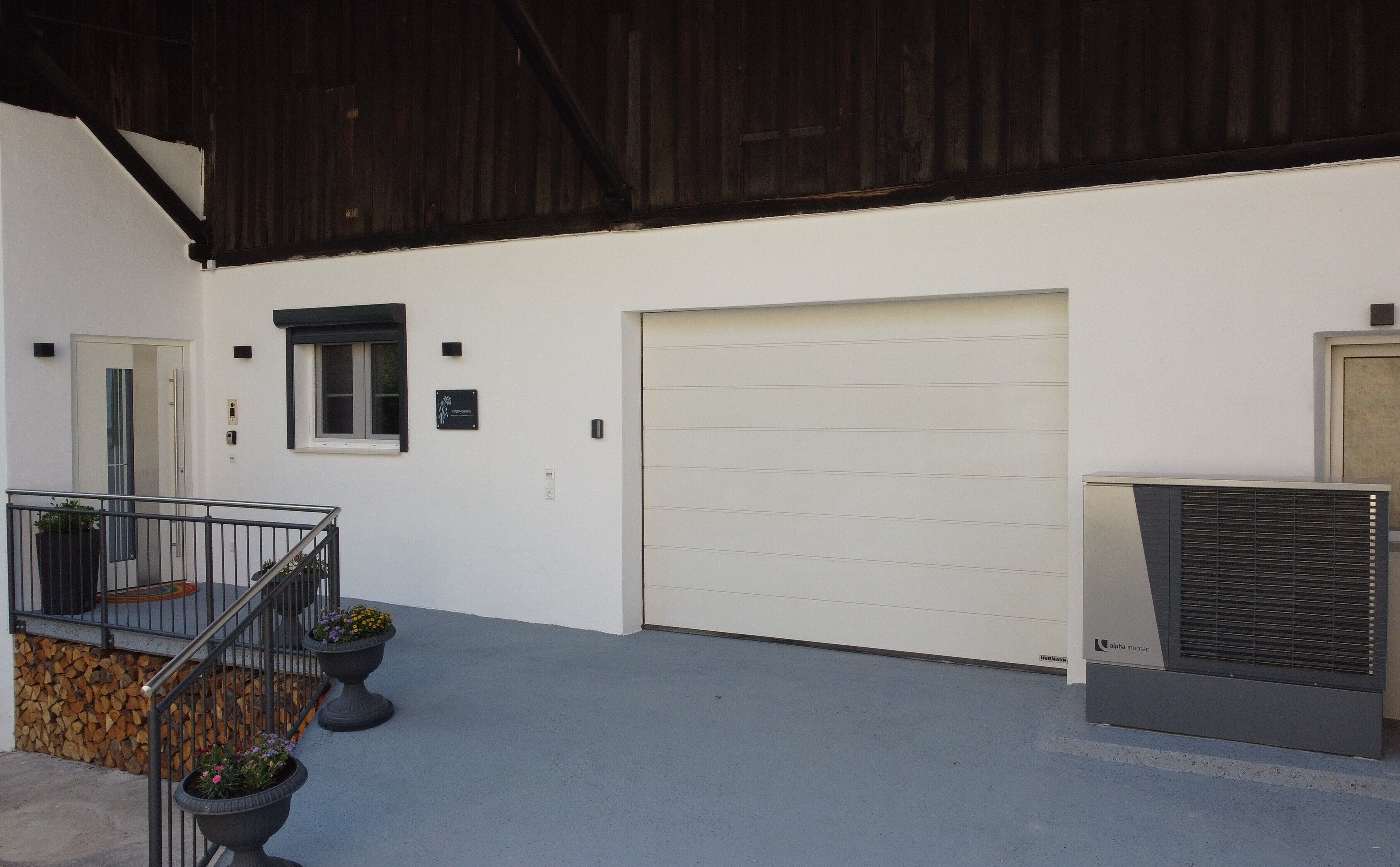 Un edificio con facciata bianca, un portone garage chiuso, una scala metallica con piante in vaso e un grande condizionatore d'aria.