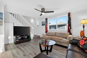 A living room with a sofa, a coffee table, a TV, and a staircase. - 1 Block To Beach • Walk To Shops + Dining | Pet Friendly | Poinciana (Lauderdale-by-the-Sea)