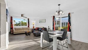 A modern living room with a sofa, coffee table, and dining area with chairs. - 1 Block To Beach • Walk To Shops + Dining | Pet Friendly | Poinciana (Lauderdale-by-the-Sea)