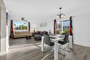 A modern living room with a sofa, coffee table, and dining area with chairs. - 1 Block To Beach • Walk To Shops + Dining | Pet Friendly | Poinciana (Lauderdale-by-the-Sea)