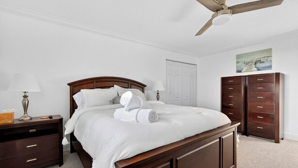 A neatly made bed with white linens and a rolled towel, a wooden nightstand with a lamp, a dresser with drawers, a ceiling fan, and a door in the background. - 1 Block To Beach • Walk To Shops + Dining | Pet Friendly | Poinciana (Lauderdale-by-the-Sea)