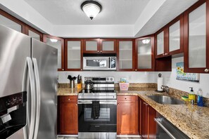 A modern kitchen with stainless steel appliances, wooden cabinets, granite countertops, and a microwave above the stove. - 1 Block To Beach • Walk To Shops + Dining | Pet Friendly | Poinciana (Lauderdale-by-the-Sea)