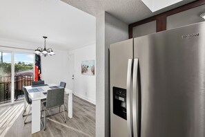 A modern kitchen with a stainless steel Whirlpool refrigerator, a dining area with a white table and grey chairs, and a balcony with a view. - 1 Block To Beach • Walk To Shops + Dining | Pet Friendly | Poinciana (Lauderdale-by-the-Sea)