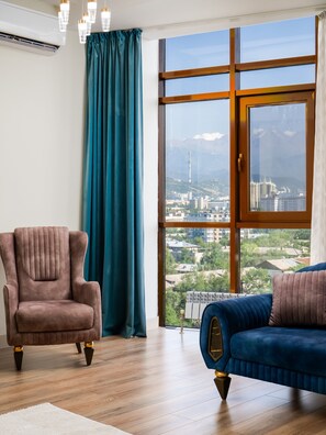 A room with a large window, a brown armchair, a blue sofa, and a wooden floor. - Spacious 4-room apartment (Almaty)
