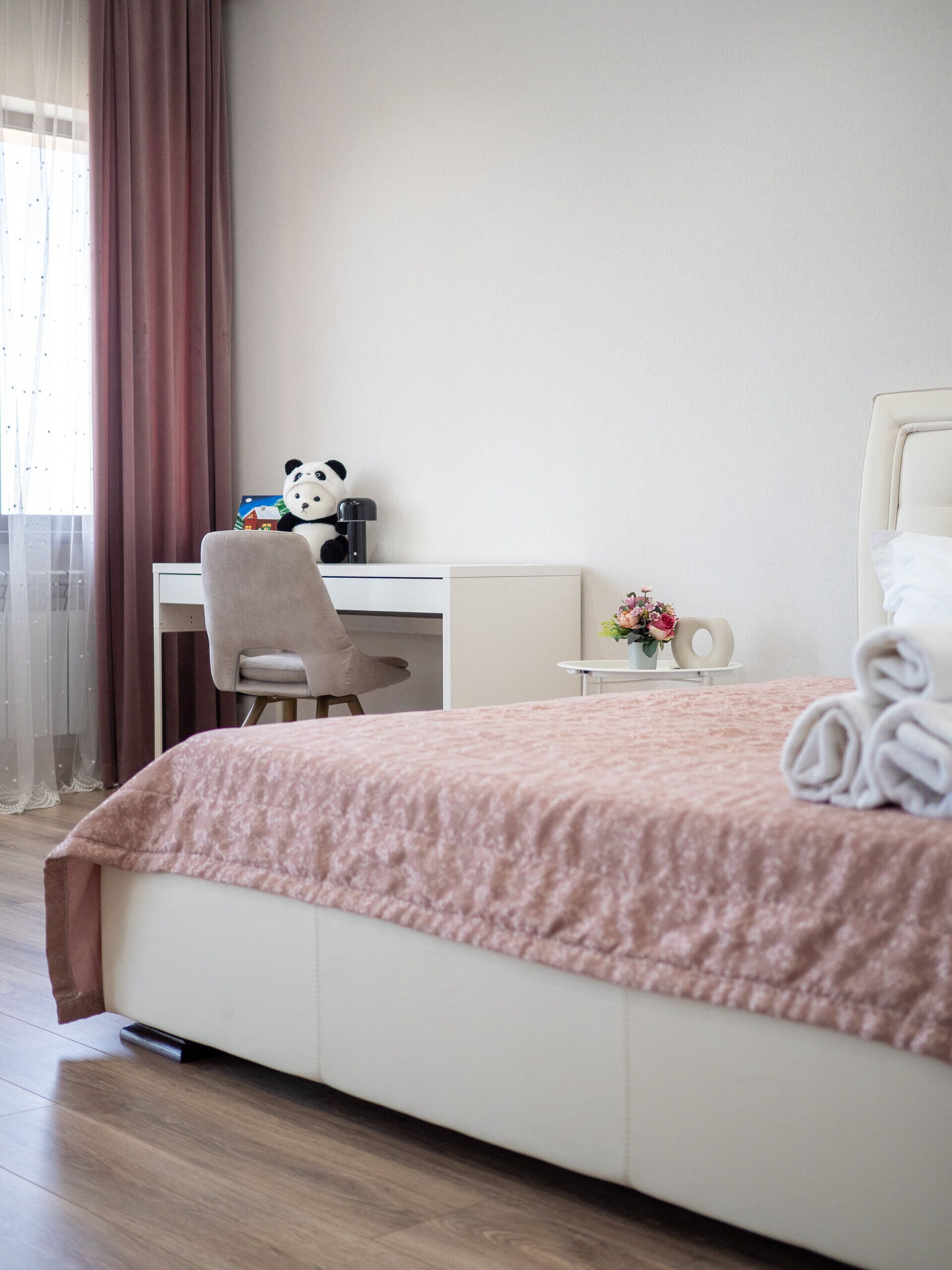 A hotel room with a bed, a desk, a chair, a panda plush toy, a vase with flowers, and a window with curtains.