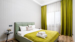 Ein modernes Hotelzimmer mit einem grünen Bett, weißer Bettwäsche, einem kleinen Nachttisch mit einer Vase und Blick durch leichte Vorhänge.