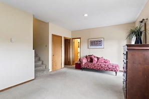 Ein Zimmer mit Treppe, einer Holztür, einem roten Ottomanen, einem gemusterten Sofa und einem gerahmten Bild an der Wand.