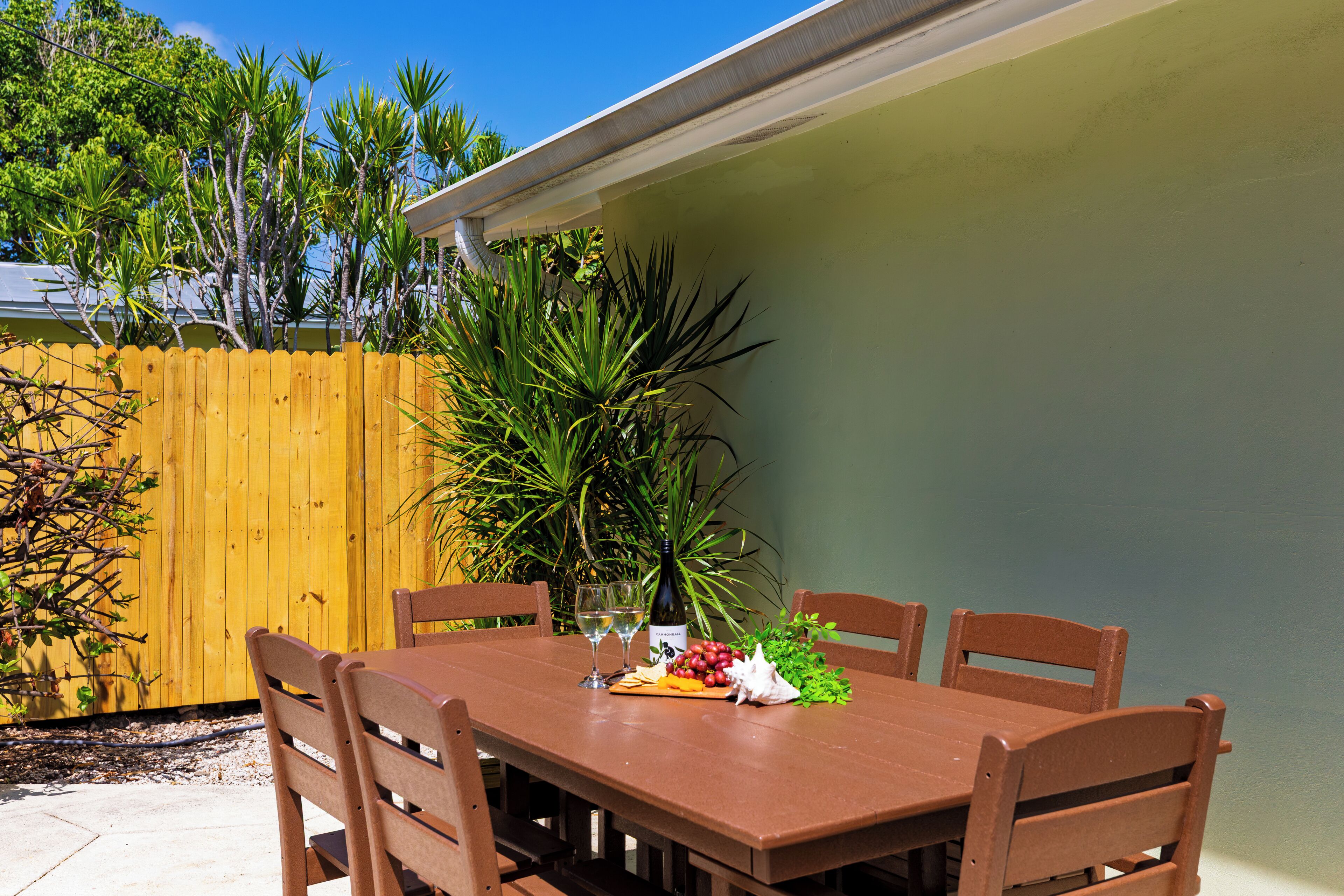 An outdoor patio with a wooden table and chairs, surrounded by a wooden fence and greenery.