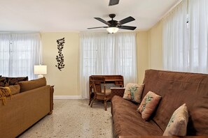 A living room with a brown sofa, a wooden chair, a ceiling fan, and sheer curtains. - Buttonwood Corner | Midtown Key West • Pool + Tropical Patio | Near Beaches (Key West)