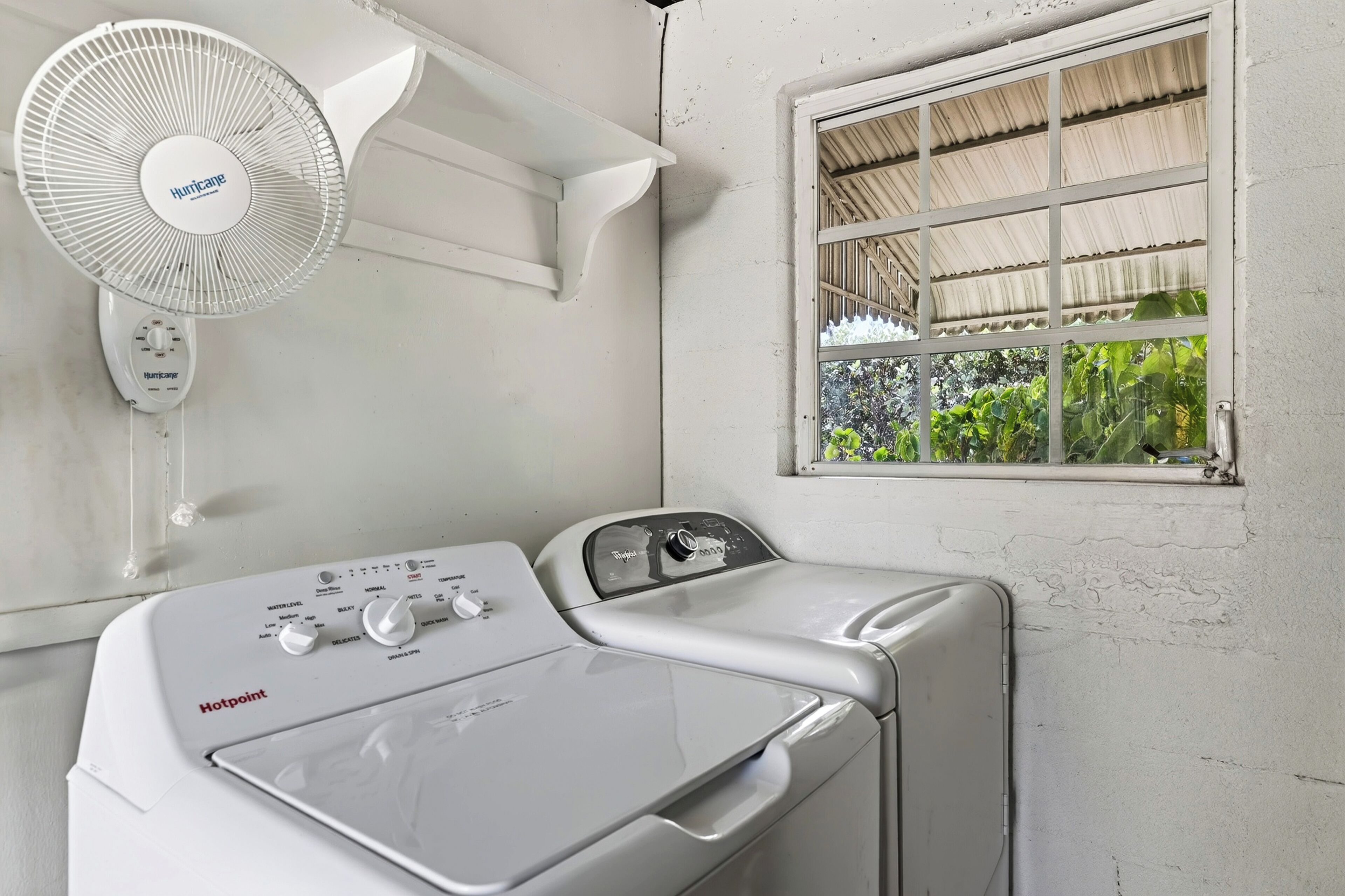 A laundry area with a white wall-mounted fan, a window with a view of greenery, and a front-loading washing machine.