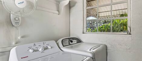 A laundry area with a white wall-mounted fan, a window with a view of greenery, and a front-loading washing machine.