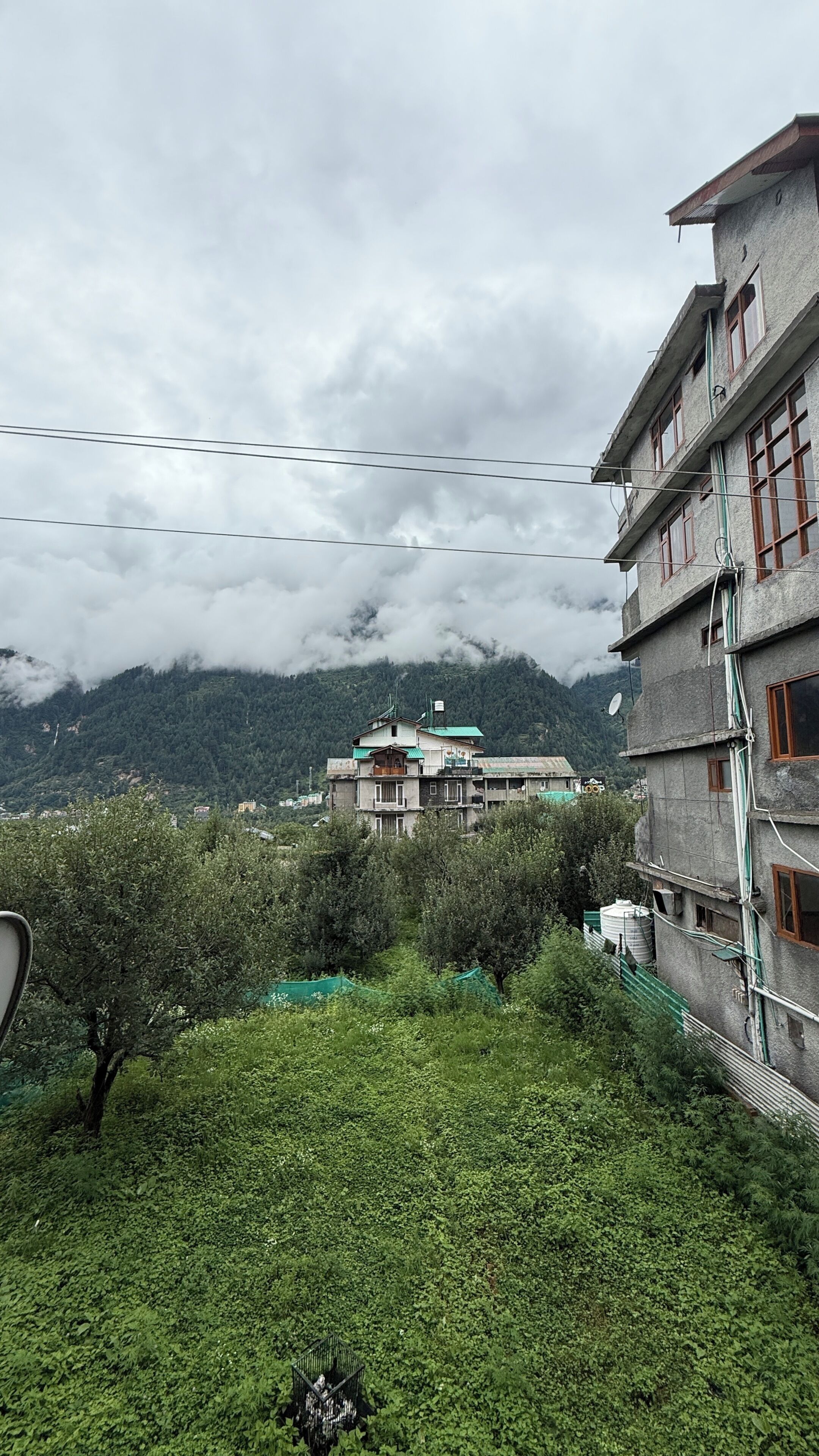 A multi-story building with a balcony and a satellite dish, surrounded by greenery and mountains in the background.