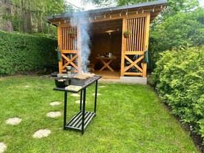 Eine Grillstation im Freien aus Holz mit Tisch und Grill, umgeben von einem gepflegten Rasen und einer Holzkonstruktion im Hintergrund.