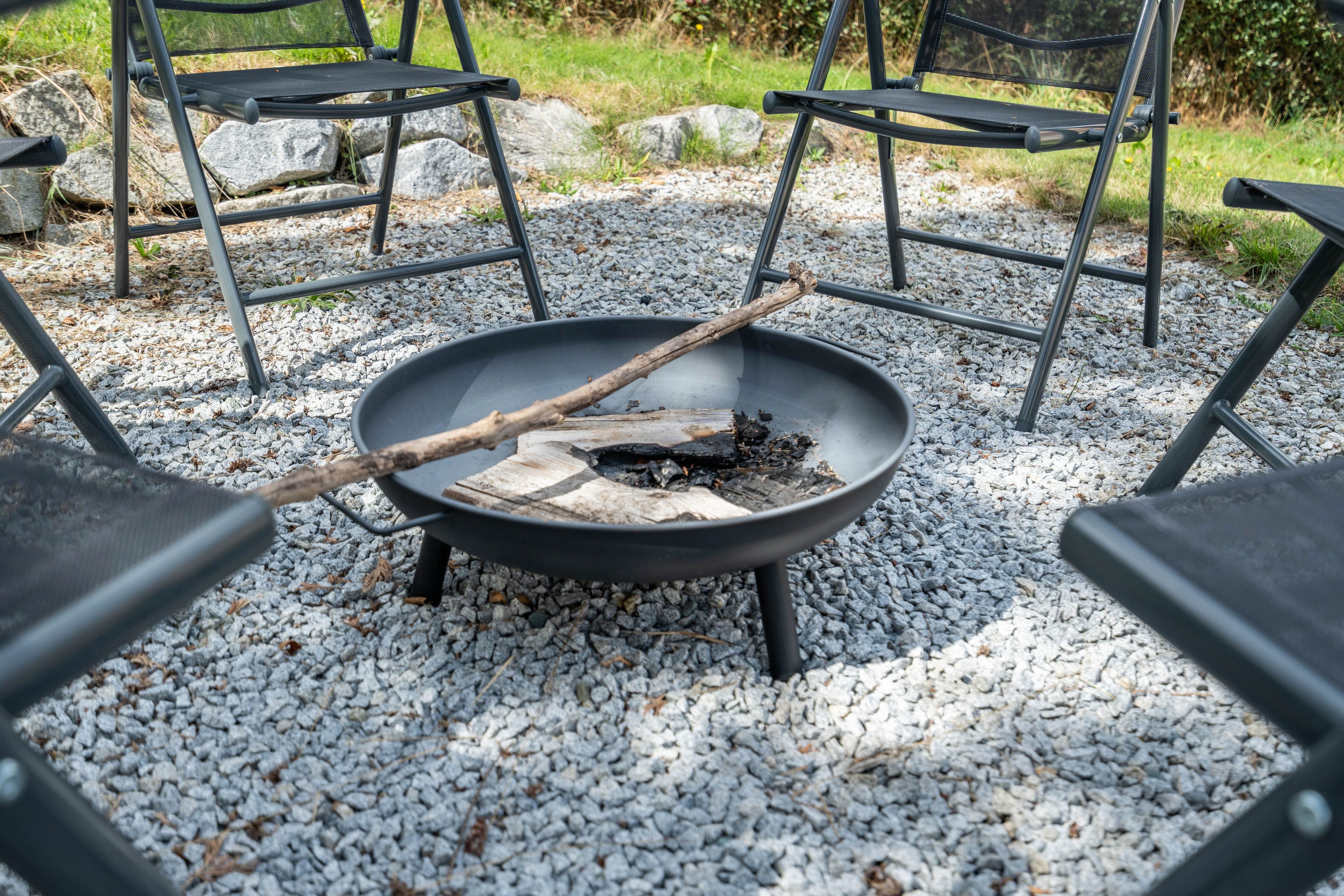 A black metal fire pit with a stick and wood pieces, surrounded by black chairs on a gravel surface.