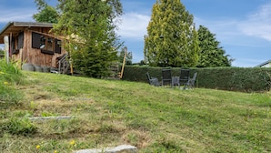 Eine Holzhütte mit einer Treppe, die zu einer Terrasse führt, umgeben von einer Rasenfläche mit vereinzelten Steinen und einer Hecke.