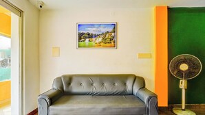 Een grijze bank in een kamer met een inlijsting foto aan de muur, een staande ventilator en een groene wand.