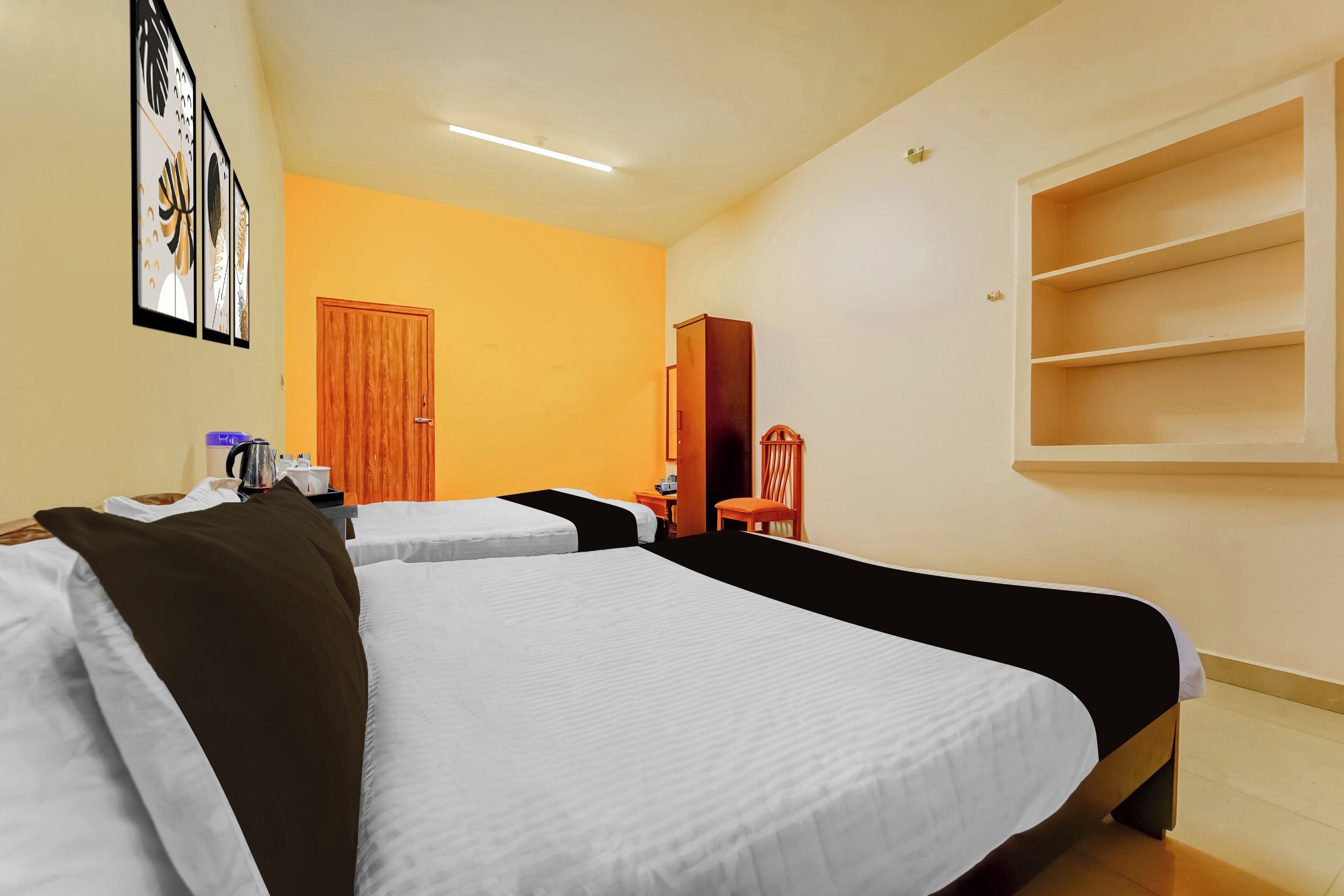 Quarto de hotel com duas camas, uma porta de madeira, uma mesinha com chaleira e uma estante.