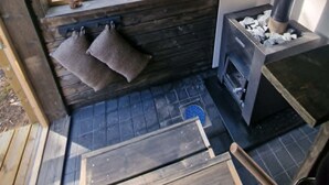 Interior de una cabaña con un ventanal grande con vistas a un bosque, una chimenea y una pequeña zona de estar con cojines.
