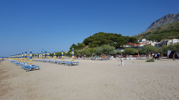 Una playa con hileras de sombrillas azules y amarillas, cielo despejado y montañas al fondo.