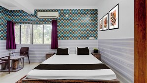 Habitación de hotel con una cama grande, un escritorio y sillas. Hay una ventana con cortinas, un aire acondicionado instalado en la pared y cuadros en la pared.