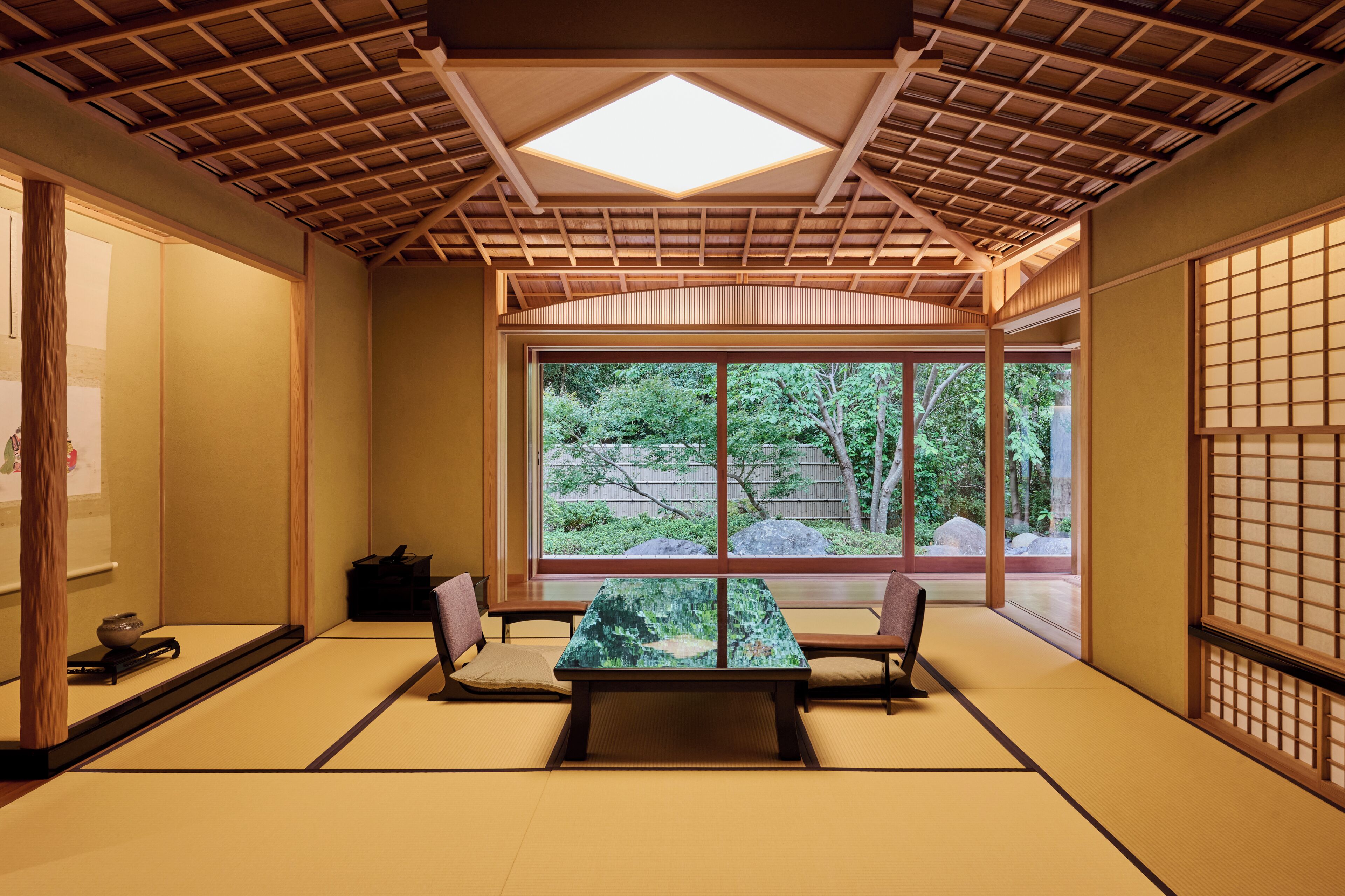 Una habitación con esteras de tatami, una mesa baja, sillas, una puerta corredera y vistas a la vegetación exterior.