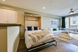 A modern bedroom with a bed, a sofa, and a view of a building through a window. - Steps to the Sand! Luxury Oceanside Pier Condo (Oceanside)