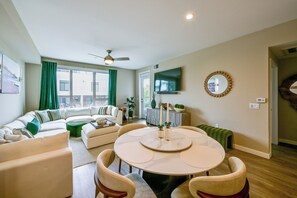 A modern living room with a sectional sofa, a dining table with chairs, a flat-screen TV mounted on the wall, and a ceiling fan. - Steps to the Sand! Luxury Oceanside Pier Condo (Oceanside)