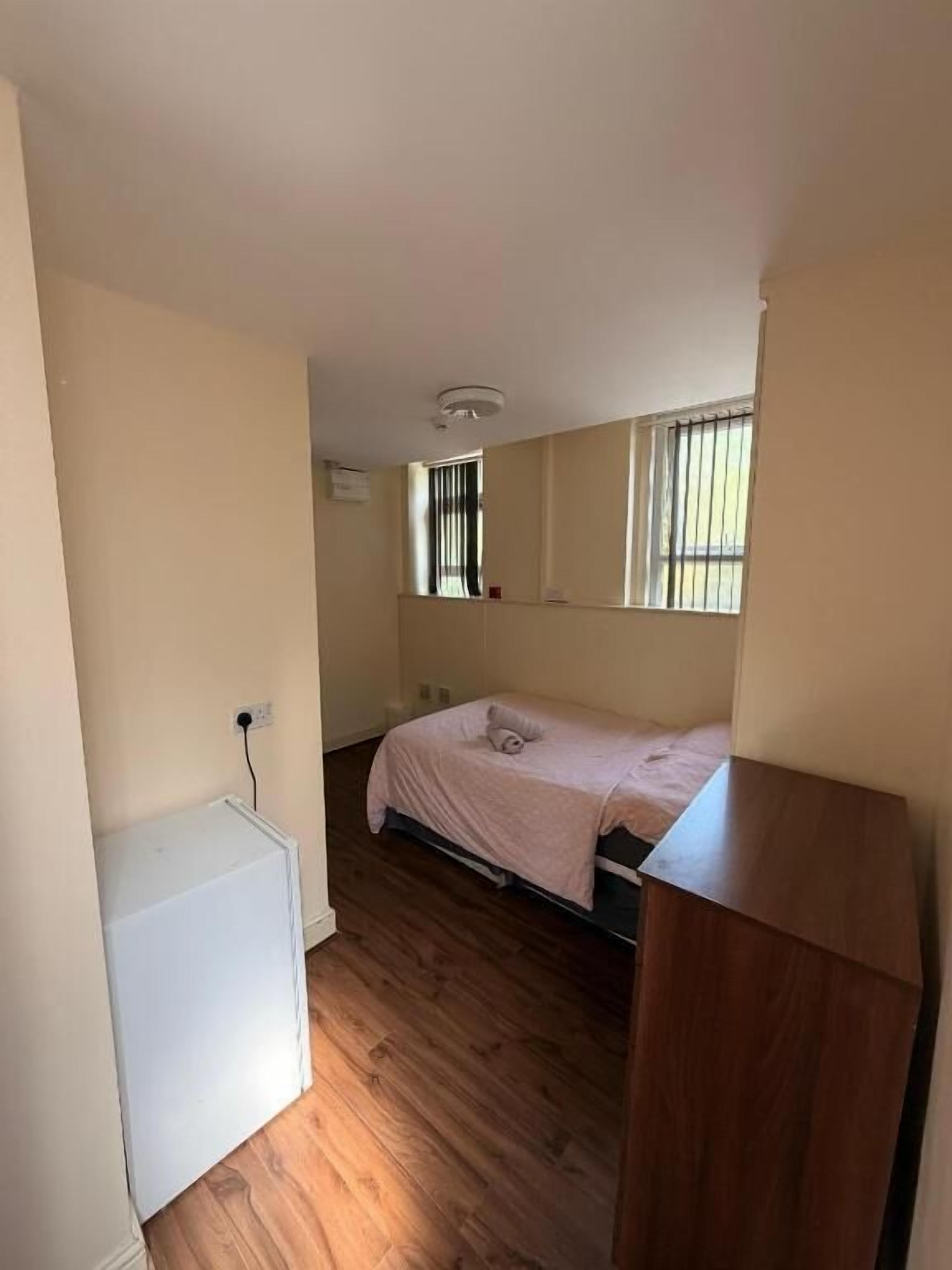 A small, single-room apartment with a bed, a wooden floor, a white refrigerator, and a window with blinds.