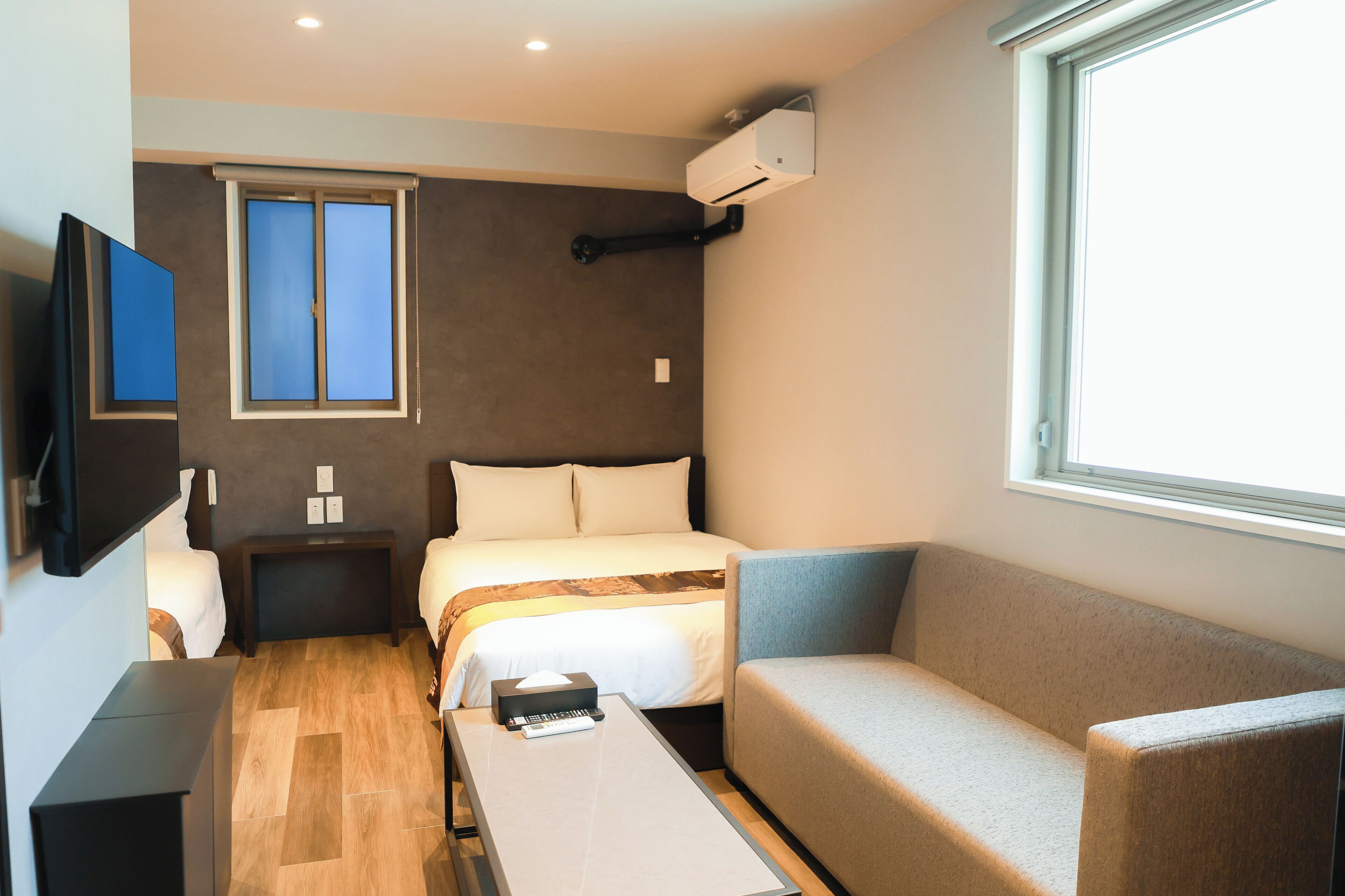 Habitación de hotel compacta con cama, sofá, televisión, ventana y aire acondicionado.
