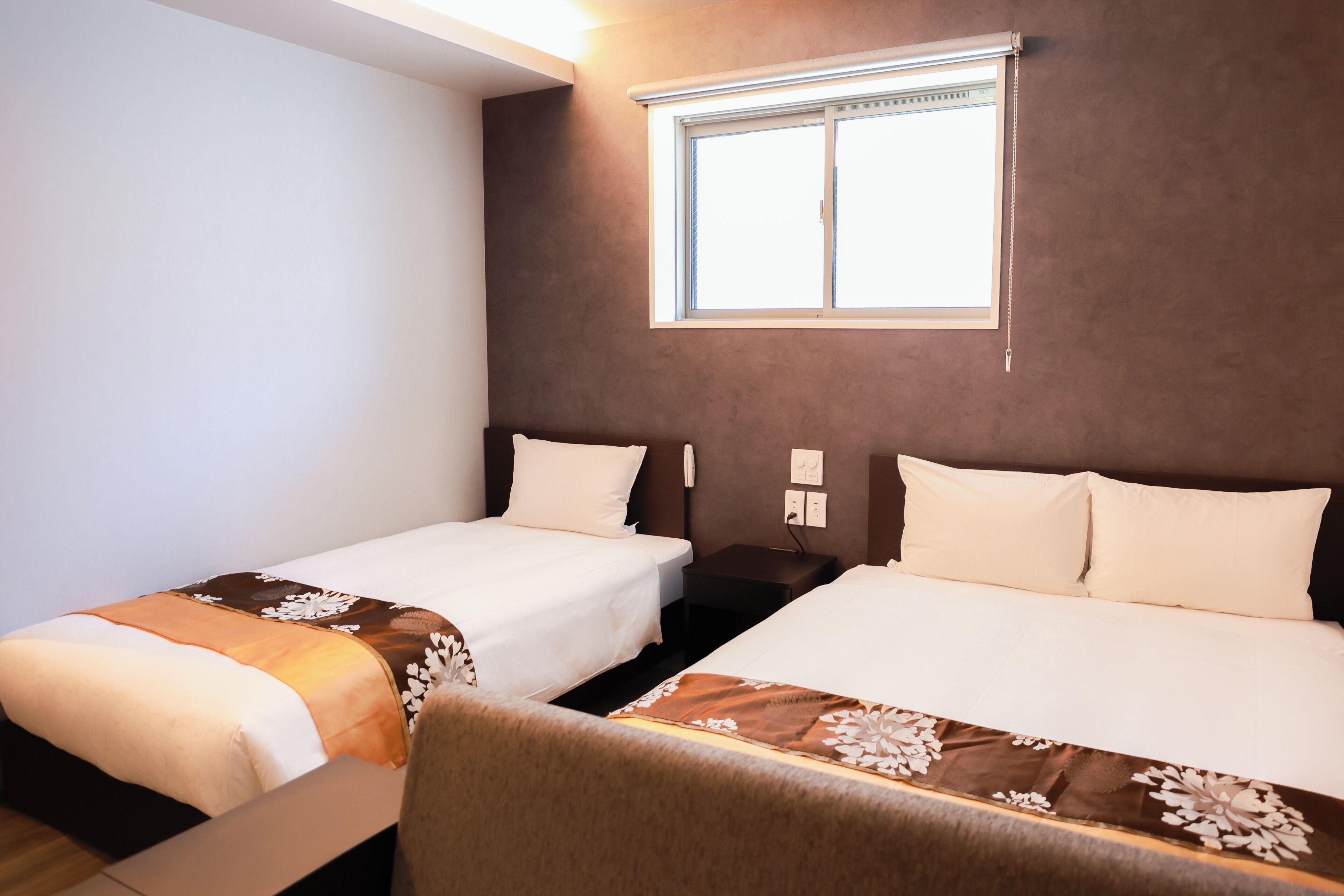 A hotel room with two beds, a window with blinds, a bedside table with a lamp, and a wall-mounted phone.