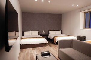 A hotel room with two beds, a sofa, a small table with a remote control, and a window with blinds.