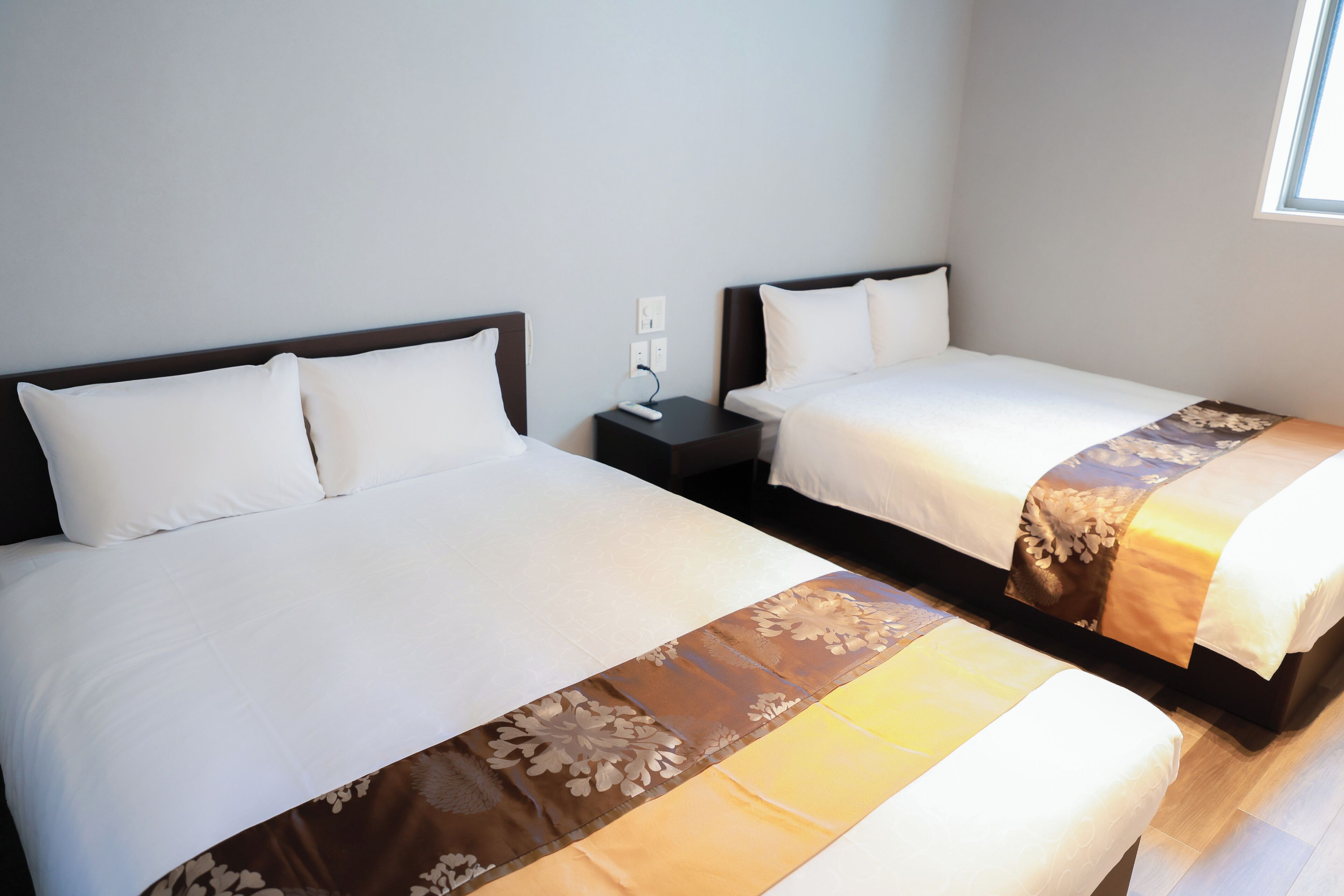 A hotel room with two beds, each with a white bedspread and a brown decorative runner. There is a nightstand with a lamp between the beds.