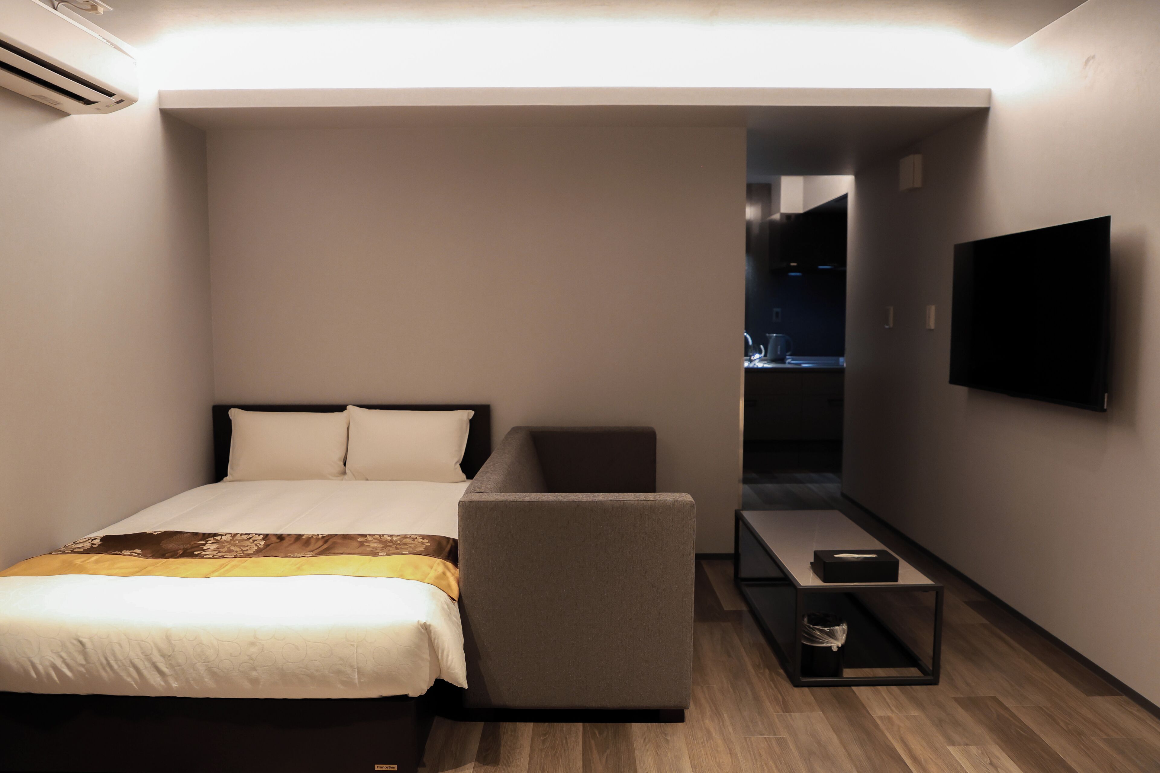 A compact hotel room with a bed, a seating area, a small table, and a TV mounted on the wall.