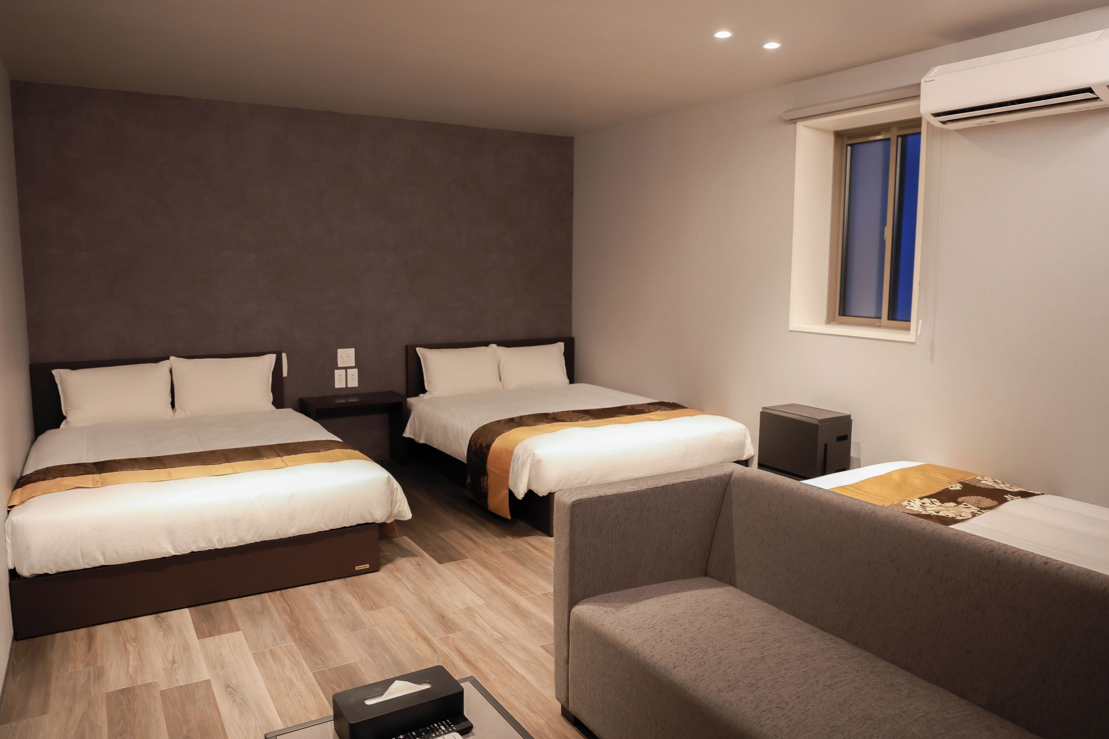 Habitación de hotel con dos camas, un sofá, una mesita de noche, un equipo de aire acondicionado y una ventana.