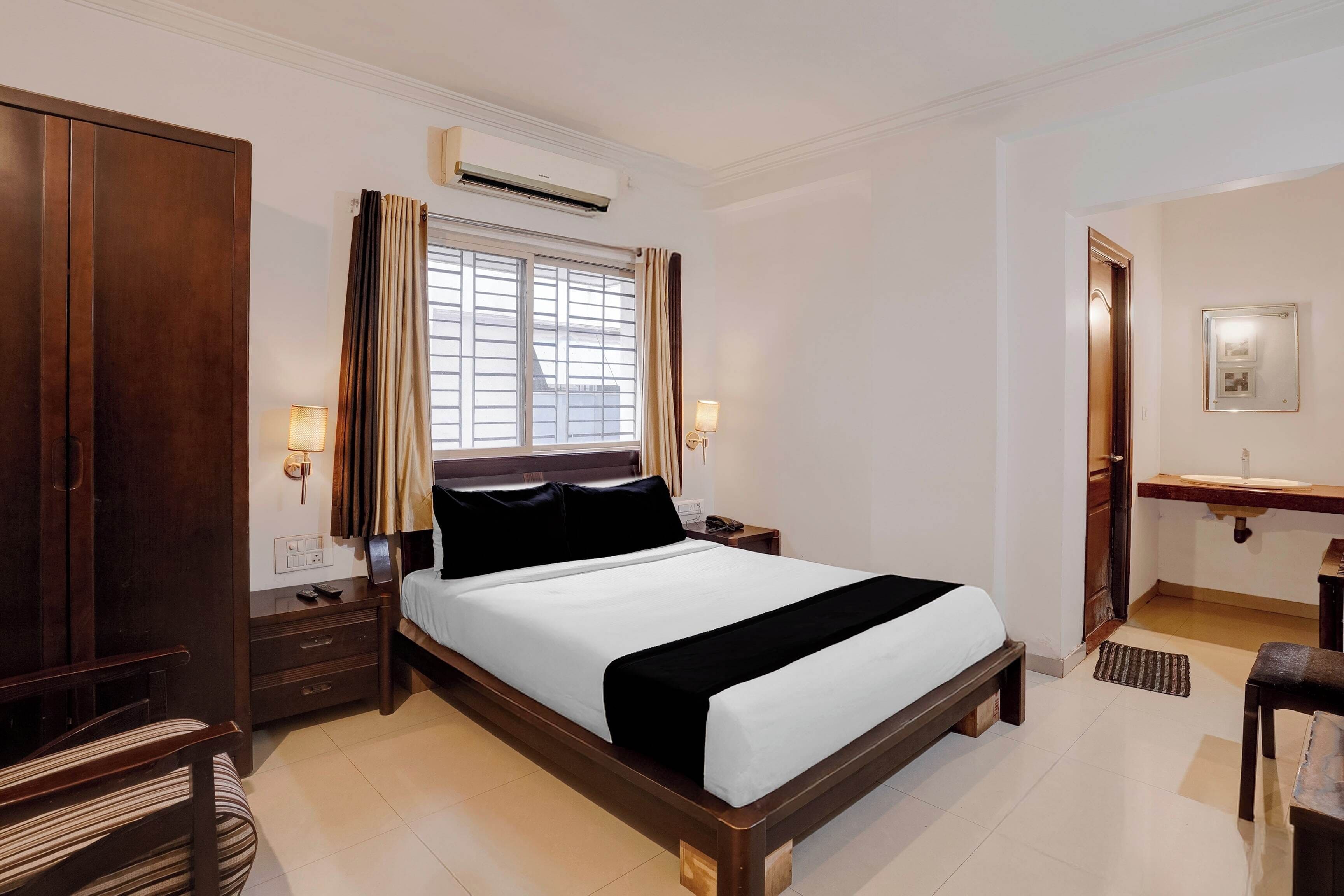 A hotel room with a large bed, a bedside table, a nightstand, a wardrobe, an air conditioning unit, a window with curtains, a bedside lamp, and a bathroom with a mirror and sink.
