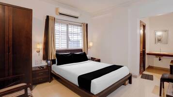 A hotel room with a large bed, a bedside table, a nightstand, a wardrobe, an air conditioning unit, a window with curtains, a bedside lamp, and a bathroom with a mirror and sink.