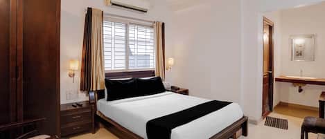 A hotel room with a large bed, a bedside table, a nightstand, a wardrobe, an air conditioning unit, a window with curtains, a bedside lamp, and a bathroom with a mirror and sink.