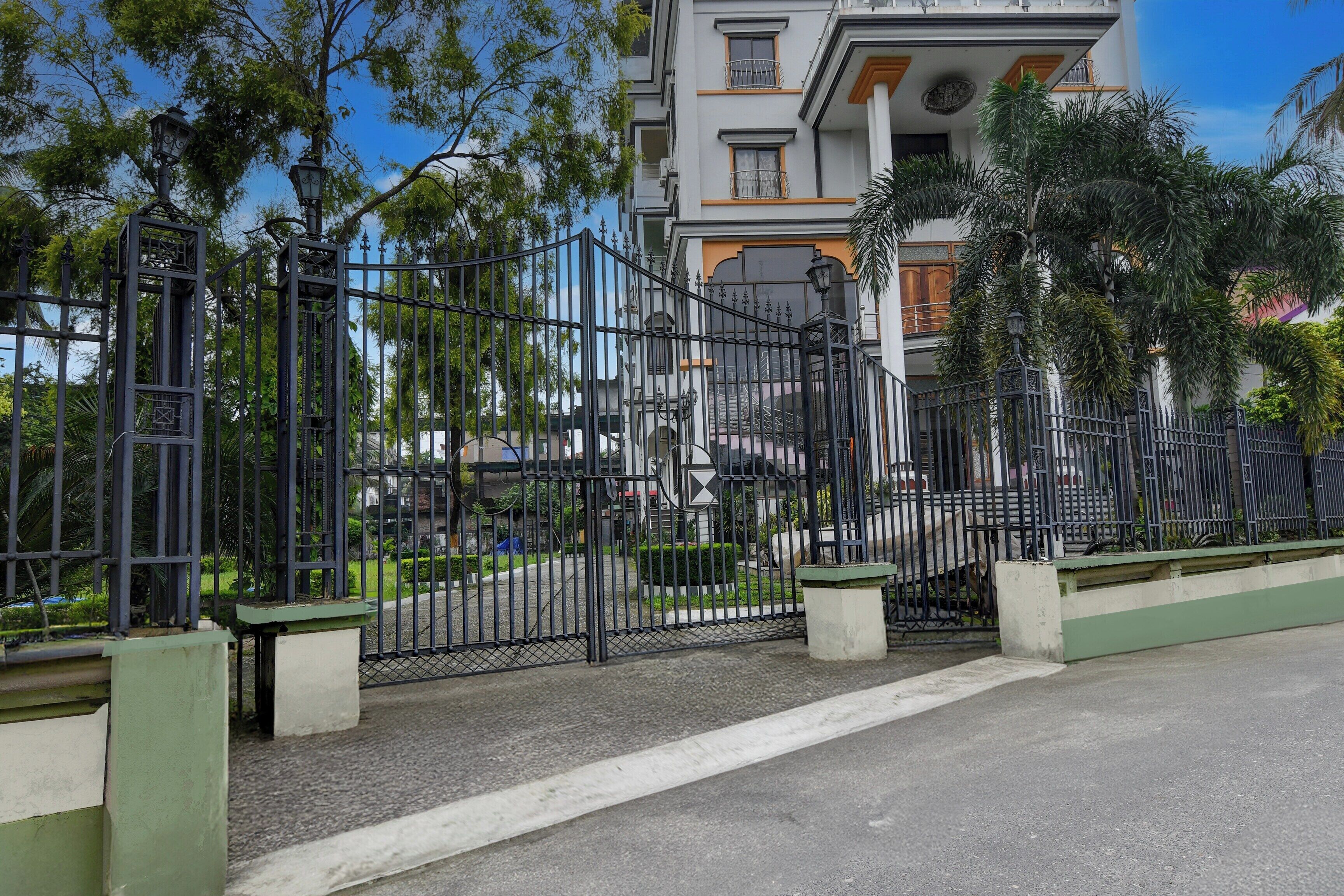 A gated entrance with a metal fence, a paved road, and a multi-story building with balconies in the background.