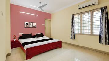 A bedroom with a bed, a ceiling fan, an air conditioner, a window with curtains, and a wall with framed pictures.