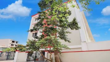 A multi-story building with a white facade and red trim, surrounded by greenery and a gated entrance.