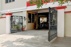 Una entrada porticada a un edificio con techo rojo, una planta en maceta y una verja metálica con un emblema de la letra 'A'.