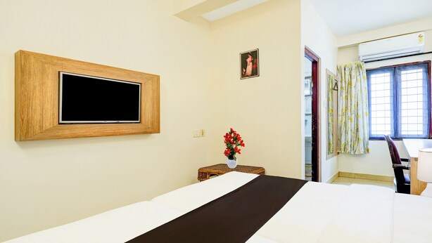 A hotel room with a bed, a television mounted on the wall, a small table with a vase of flowers, and a window with curtains. - Super Collection O MG Road (Kanayannur)