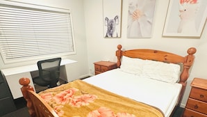 Un dormitorio con cama de madera, un escritorio y una silla. Se observan obras de arte en las paredes y una ventana con persianas.