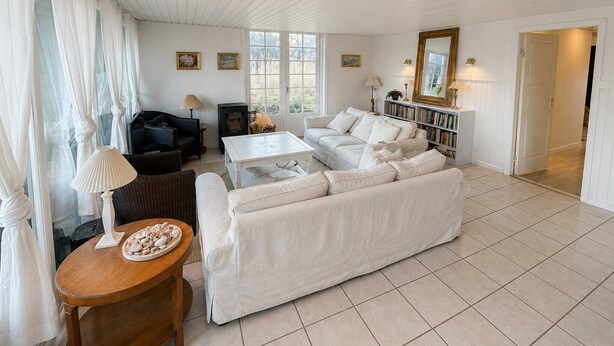 Ampla sala de estar com sofá branco, mesa de centro e lareira. Há estantes de livros e uma porta que dá para outro quarto.