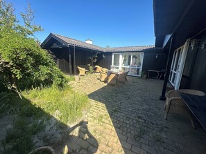 Ein überdachter Außenbereich mit Pflasterpatio, Korbmöbeln und einem Gebäude mit Satteldach.