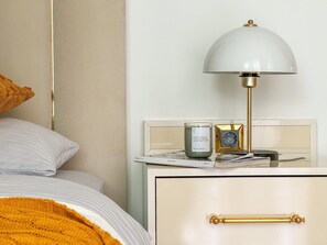 A neatly made bed with a white and gold color scheme, a bedside lamp, a small table with a clock and a candle, and a drawer with a gold handle. - Qoople Legend 36 В-04 100 Meter vom Strand entfernt by Interhome (Alanya)