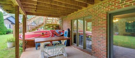 A covered patio area with a wooden table and chairs, a red boat, and a brick wall with large windows.