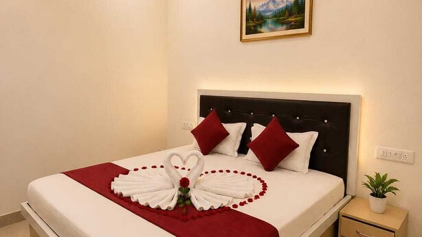 A neatly made bed with a heart-shaped towel arrangement and red pillows, a small bedside table with a plant, and a painting on the wall. - KU Residency (Varanasi)