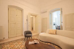 A living room with a sofa, a chair, a coffee table, and a patterned rug. There are white doors and an air conditioning unit on the wall. - TD Palazzo Fiume 1 Heart of Polignano (Polignano a Mare)