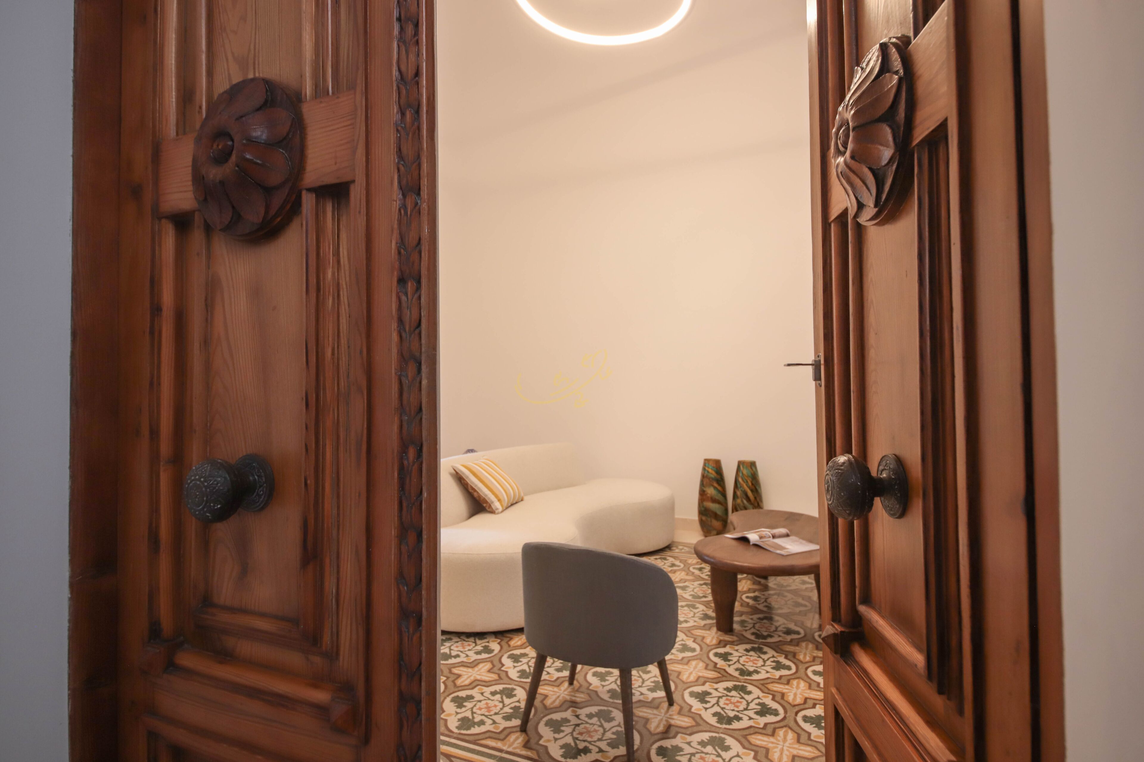 Uma porta de madeira com maçaneta entalhada, um sofá branco com almofada estampada, uma cadeira cinza e uma pequena mesa com um vaso.